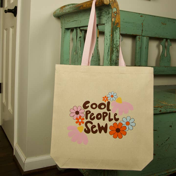 Beige tote bag with 'Cool People Sew' text and floral designs hanging on a green wooden chair.