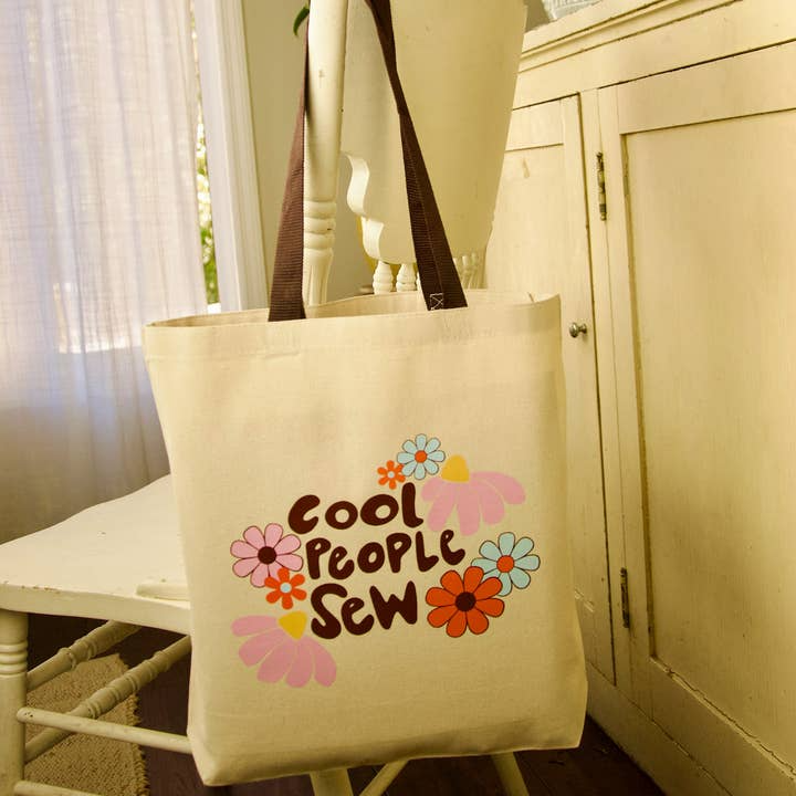 Beige tote bag with 'Cool People Sew' text and floral designs on a chair.