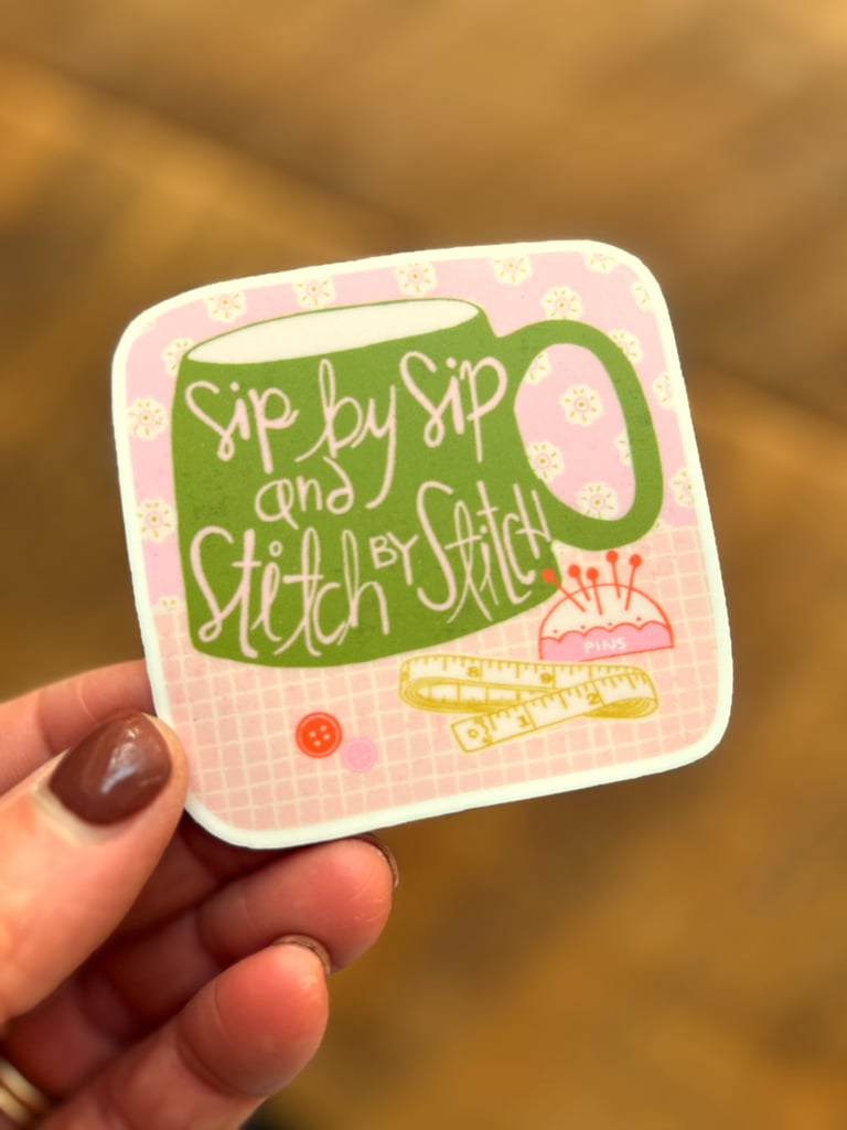 Sticker with 'Sip by Sip and Stitch by Stitch' text held by a hand on a blurred background