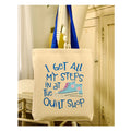Beige tote bag with blue text and shoe graphic on a mannequin in a quilt shop setting.