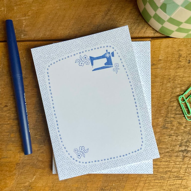 Two white cards with blue sewing machine design on a wooden surface with a pen and paper clips.