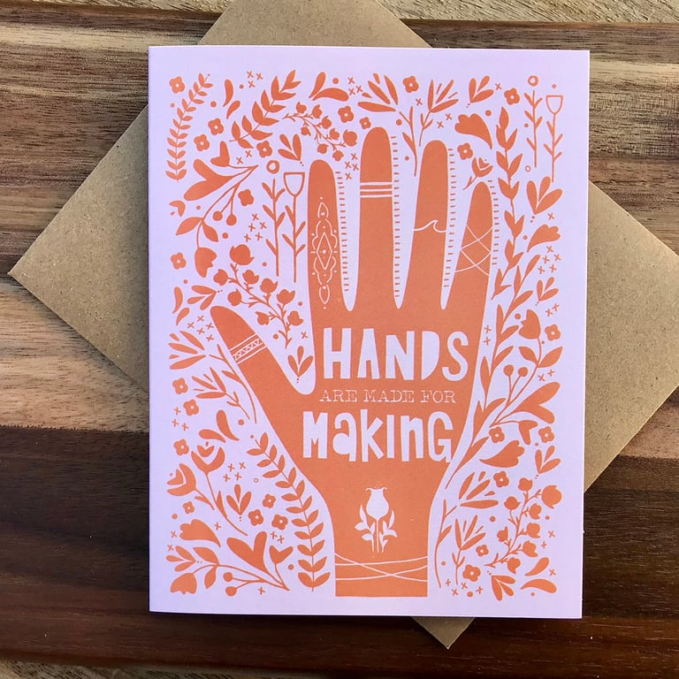 Greeting card with orange hand design and 'Hands are made for making' text on a wooden surface.