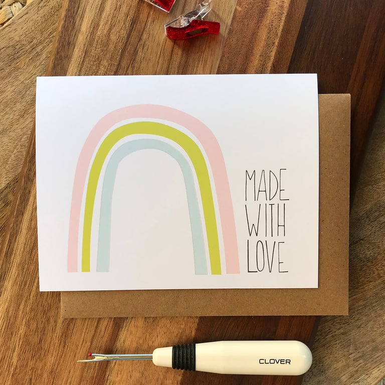 Card with rainbow design and 'Made with Love' text on a wooden surface with a sewing tool.