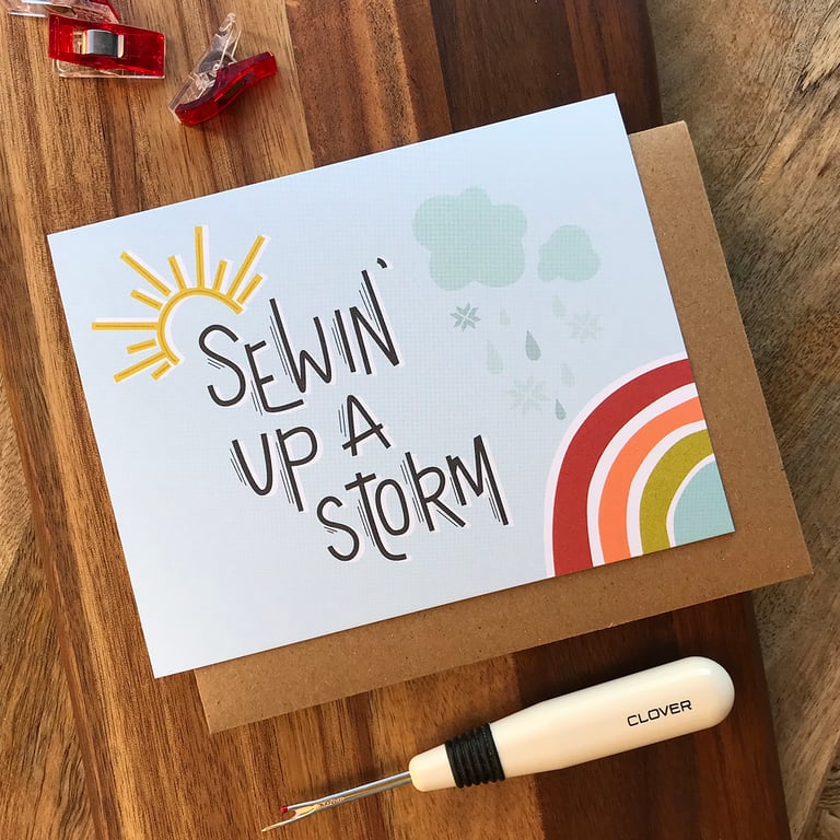 Card with 'Selvin' Up A Storm' text, rainbow, and sun on a wooden surface with a sewing tool.
