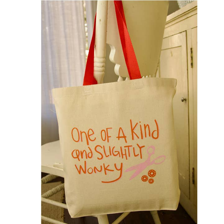 Beige tote bag with red handles and text 'One of a Kind and Slightly Wonky' on a chair.
