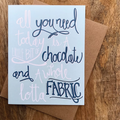 Card with text 'All you need today is a little bit of chocolate and a whole lotta fabric' on a wooden surface.