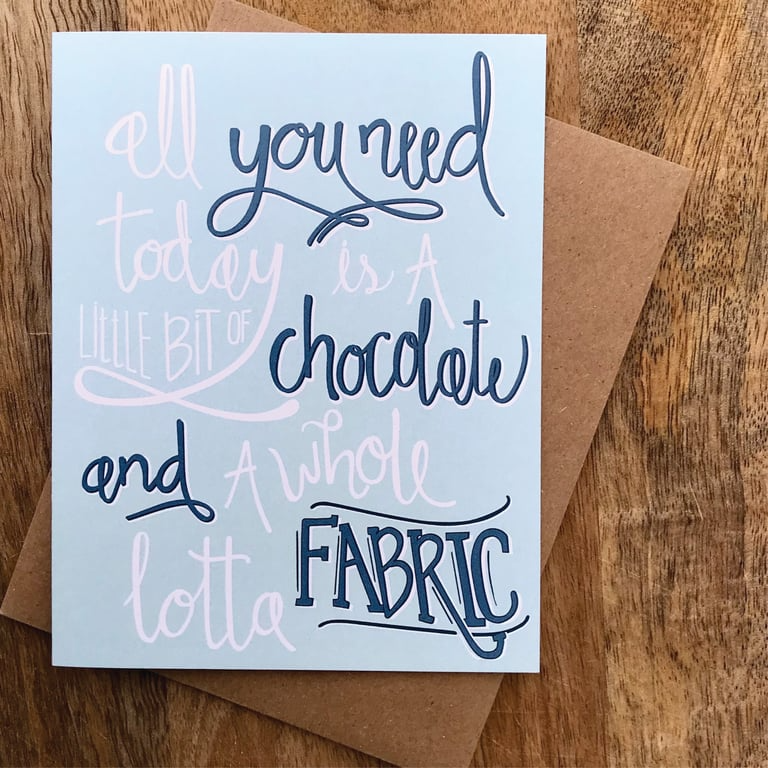 Card with text 'All you need today is a little bit of chocolate and a whole lotta fabric' on a wooden surface.